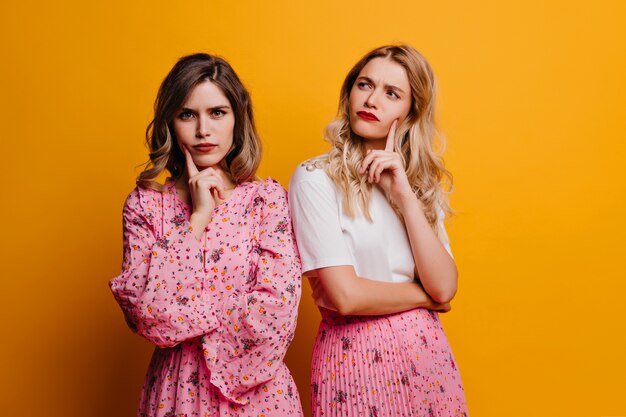 Attractive girl with red lips posing beside friend. European young ladies in pink clothes standing on yellow wall.