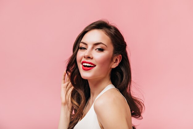 Attractive girl with red lipstick and curly dark hair looking at camera on pink background.