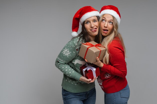 Attractive girls friends in red and white christmas hats holds a presents for each other