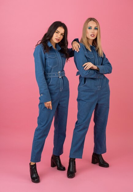 Attractive girls posing on pink background in denim overall fashion trend