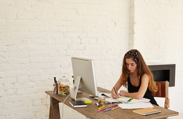 Attractive graphic designer female draws sketches of new logo for dental clinic sitting at the desk with PC computer, documents and colored stationery. Copy space wall for advertising content or text