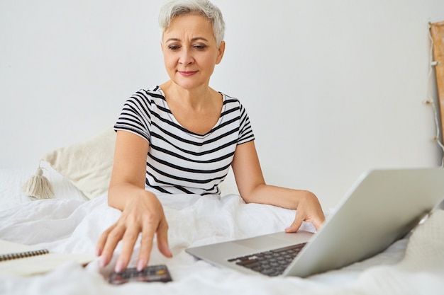 Attractive gray haired elderly businesswoman working remotely right from bedroom, sitting on bed with portable computer, using calculator, managing finances, having confident happy expression