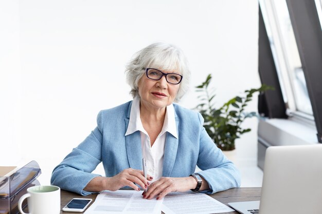 Attractive gray haired senior businesswoman in fashionable suit and glasses working in her office, sitting at desk with open laptop and papers, filling in financial documents, having serious look