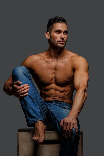 Attractive guy in blue jeans with naked torso sitting on a podium. GIsolated on grey background