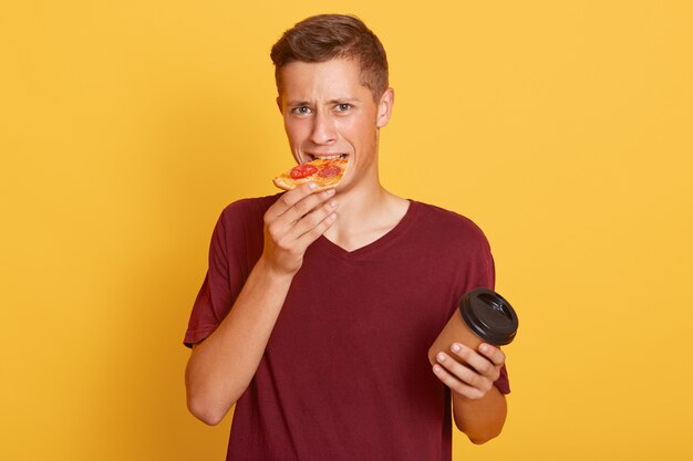 Attractive guy in casual burgundy t shirt holding hot drink in paper cup and biting slice of pizza,