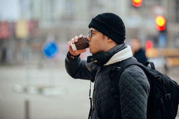 Attractive guy in eyeglasses drinking coffee, outdoors. Dressed in warm jacket and hat