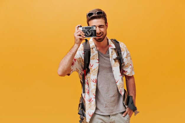 Attractive guy with cool hairstyle in grey tshirt and printed summer shirt making photo and smiling on isolated orange backdrop