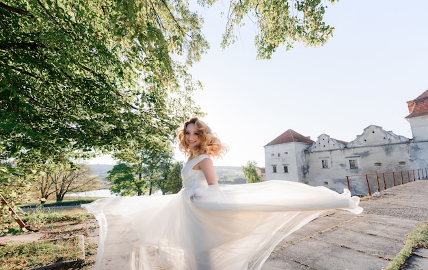 Привлекательная счастливая блондинка в белом платье оборачивается и улыбается в солнечный день возле старого каменного замка