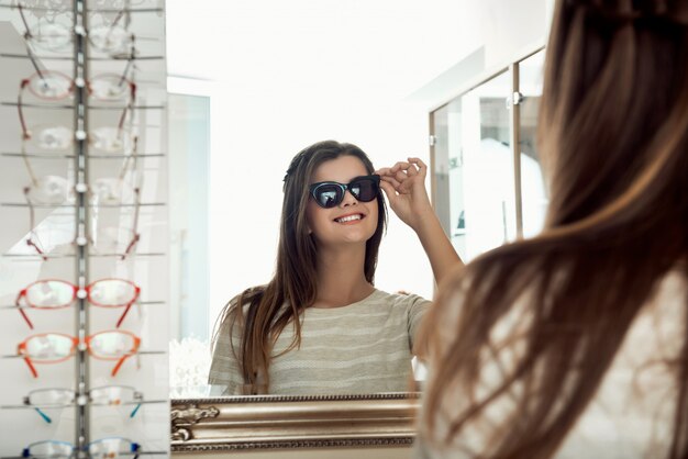 안경점에서 선글라스를 시도하는 동안 거울을보고 매력적인 행복 갈색 머리 여자