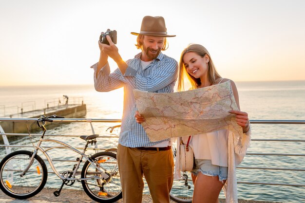 Attractive happy couple traveling in summer on bicycles, man and woman with blond hair boho hipster style fashion having fun together, looking in map sightseeing taking photos on camera