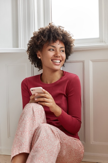 아프로 헤어 스타일을 가진 매력적인 행복한 여성 모델은 잠옷을 입고 즐거운 문자 메시지를 읽고 긍정적으로 미소 짓고 바닥에 앉아 집에서 자유 시간을 보냅니다. 화려한 여자 친구 전화를 기다립니다.