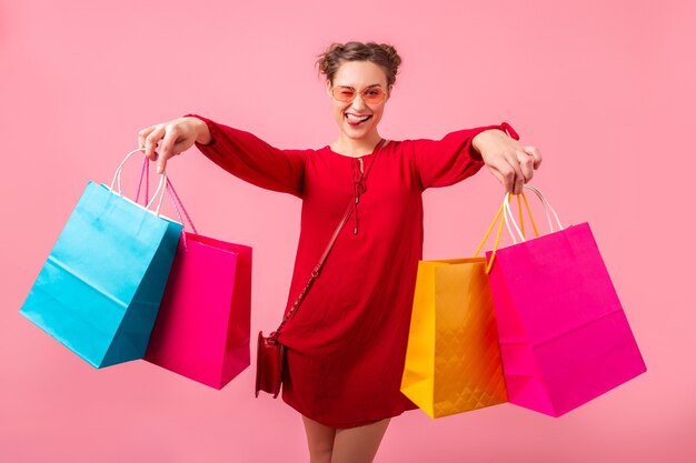 Attractive happy funny emotion stylish woman shopaholic in red trendy dress holding colorful shopping bags on pink wall isolated, sale excited, spring summer fashion trend