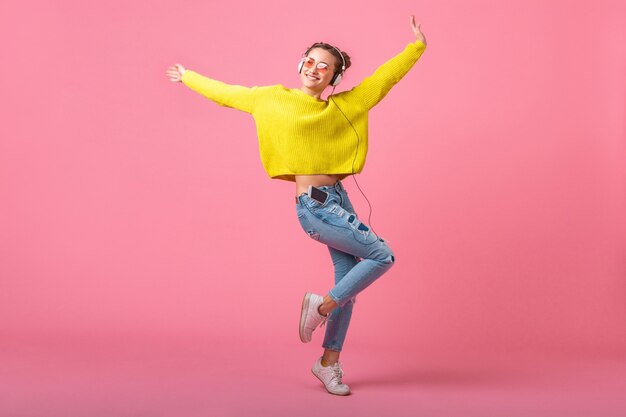 ピンクの壁に分離されたヒップスターのカラフルなスタイルの服を着て、黄色のセーターとサングラスを身に着けて、楽しんでヘッドフォンで音楽を聴いてジャンプする魅力的な幸せな面白い女性
