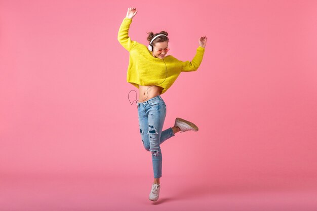 Привлекательная счастливая смешная женщина прыгает, слушая музыку в наушниках, одетая в красочный стиль хипстера, изолированного на розовой стене, в желтом свитере и солнцезащитных очках, веселится
