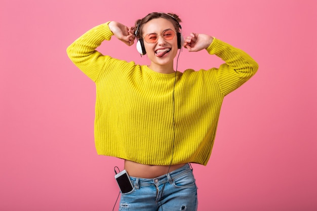 Attractive happy funny woman listening to music in headphones dressed in hipster colorful style outfit isolated on pink wall, wearing yellow sweater and sunglasses, having fun