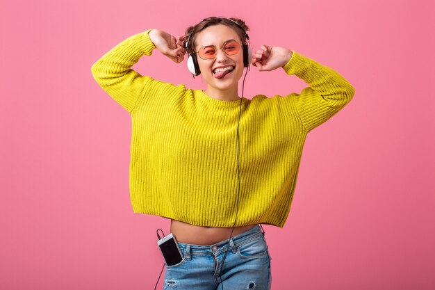 Привлекательная счастливая смешная женщина, слушающая музыку в наушниках, одетая в красочный хипстерский наряд, изолирована на розовой стене, в желтом свитере и солнцезащитных очках, веселится