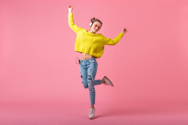 Привлекательная счастливая смешная женщина в желтом свитере прыгает, слушая музыку в наушниках, одетая в красочный хипстерский наряд, изолированный на розовой стене, весело