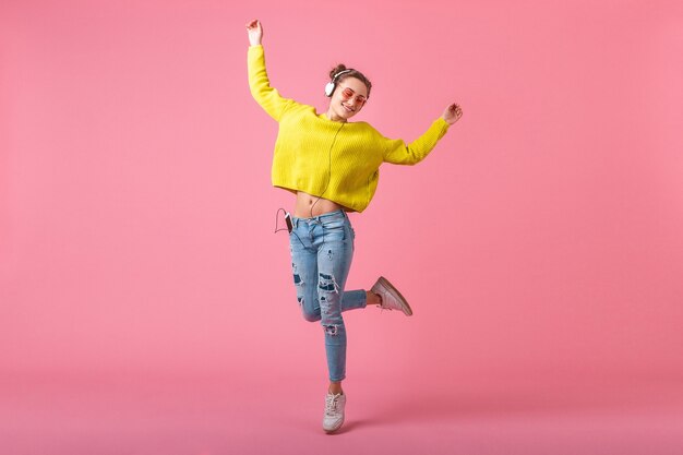 Attractive happy funny woman in yellow sweater jumping listening to music in headphones dressed in hipster colorful style outfit isolated on pink wall, having fun