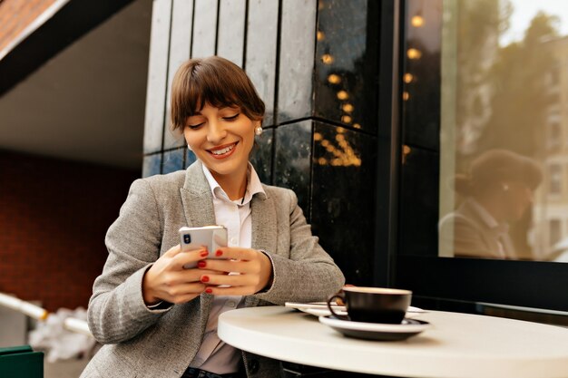 Attractive happy lady in stylish outfit using smartphone. Brunette tanned woman in grey jacket sits outside with phone, glass of coffee and laptop.