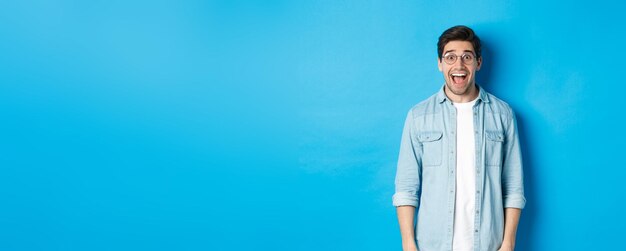 Attractive happy man in glasses looking surprised checking out advertisement standing over blue back