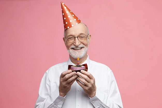 Free photo attractive happy retired caucasian male wearing bow tie, glasses and cone hat celebrating his 80th anniversary, posing isolated with birthday cake in his hands, going to blow out candle and make wish