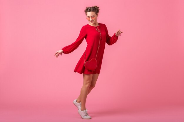 Привлекательная счастливая улыбающаяся стильная женщина в красном модном платье, прыгающая, танцующая на розовой стене, изолирована, весенне-летняя мода, романтическое настроение кокетливой девушки