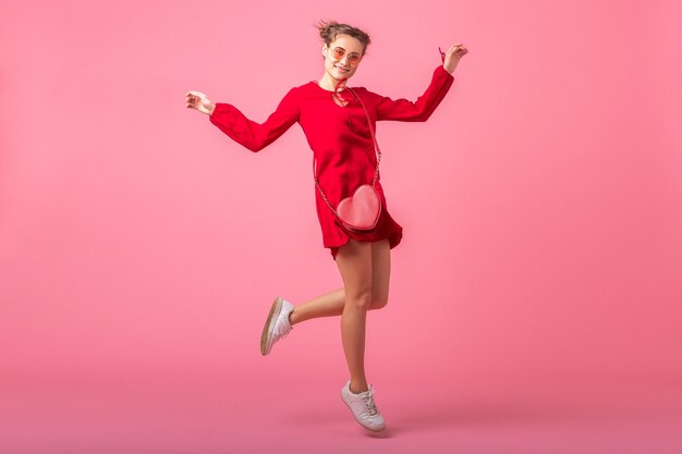 Attractive happy smiling stylish woman in red trendy dress jumping dancing on pink wall isolated, spring summer fashion trend, romantic mood flirty girl