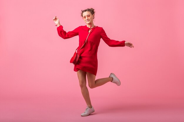 Привлекательная счастливая улыбающаяся стильная женщина в красном модном платье, прыгающая на розовой стене, изолирована, весенне-летняя мода, романтическое настроение кокетливой девушки