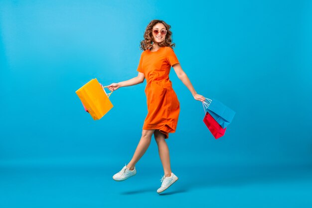 Attractive happy smiling stylish woman shopaholic in orange trendy oversize dress jumping holding shopping bags on blue studio background isolated
