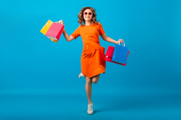 Attractive happy smiling stylish woman shopaholic in orange trendy oversize dress jumping running holding shopping bags on blue background isolated