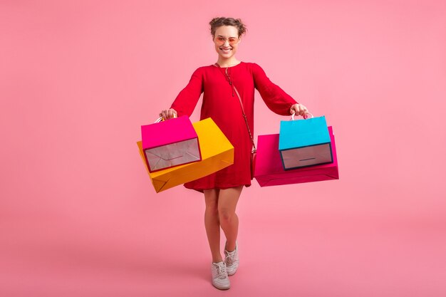ピンクの壁にカラフルなショッピングバッグを保持している赤いトレンディなドレスで買い物中毒の魅力的な幸せな笑顔のスタイリッシュな女性は、分離、販売興奮、ファッショントレンド