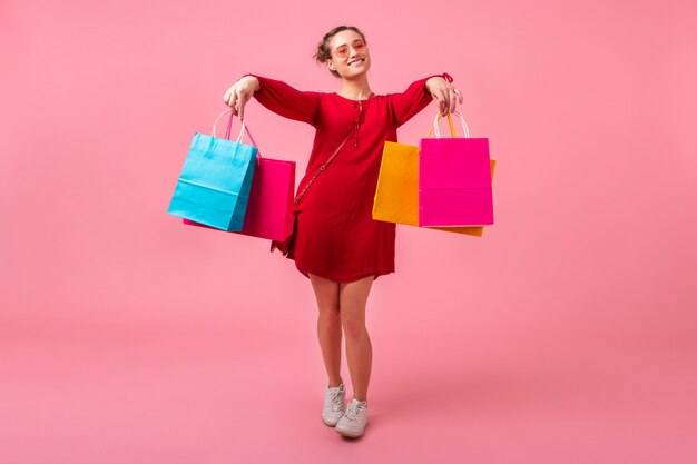 ピンクの壁にカラフルなショッピングバッグを保持している赤いトレンディなドレスで買い物中毒の魅力的な幸せな笑顔のスタイリッシュな女性は、分離、販売興奮、ファッショントレンド