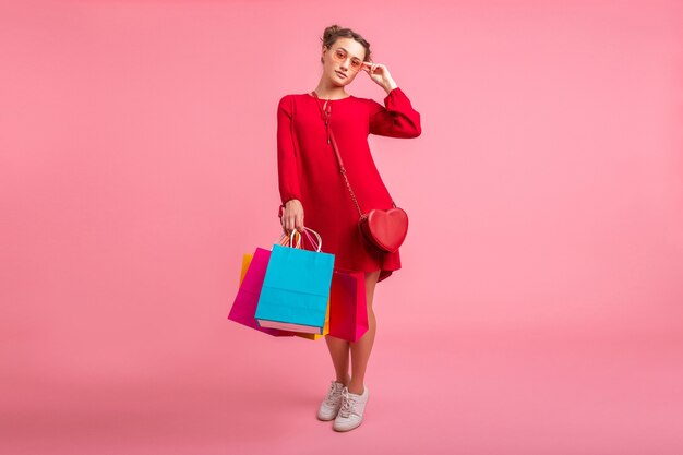 Привлекательная счастливая улыбающаяся стильная женщина-шопоголик в красном модном платье, держащая красочные сумки на розовой стене, изолированные, возбужденные продажи, весенне-летняя модная тенденция