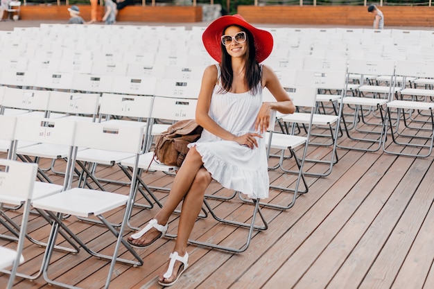 Attraente donna sorridente felice vestita in abito bianco, cappello rosso, occhiali da sole seduti in un teatro all'aperto estivo da solo, molte sedie, tendenza della moda di strada primaverile, ciao agitando la mano