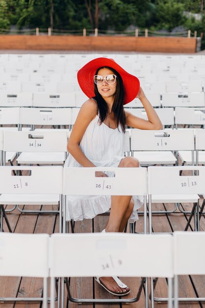 흰 드레스, 빨간 모자, 혼자 의자에 여름 야외 극장에 앉아 선글라스, 봄 거리 스타일 패션 트렌드를 입은 매력적인 행복 웃는 여자