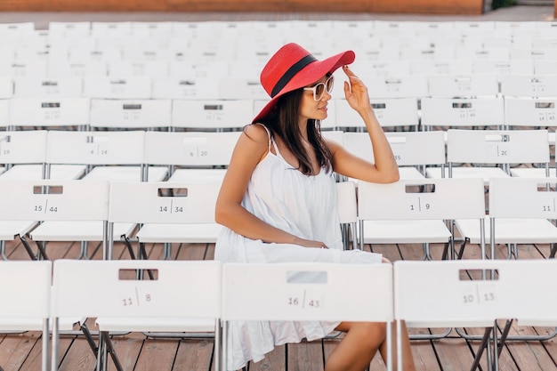 Attractive happy smiling woman dressed in white dress, red hat, sunglasses sitting in summer open air theatre on chair alone, spring street style fashion trend
