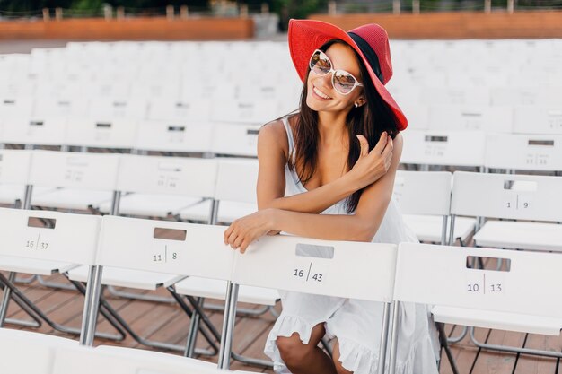 Привлекательная счастливая улыбающаяся женщина, одетая в белое платье, красную шляпу, солнцезащитные очки, сидя в летнем театре под открытым небом на стуле, весенняя уличная мода