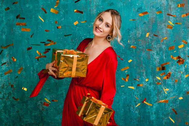 贈り物でクリスマスと新年を祝うスタイリッシュな赤いドレスの魅力的な幸せな笑顔の女性