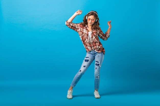 Attractive happy woman posing in cheerful mood listening to music in headphones in checkered shirt and jeans isolated on blue studio background, looking up