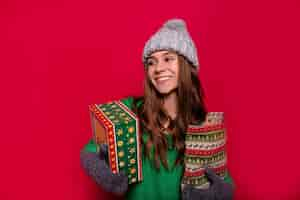 Free photo attractive happy woman with long light-brown hair and wonderful smile dressed winter grey cap, mittens and green sweater holding new year presents and smiling on isolated red background