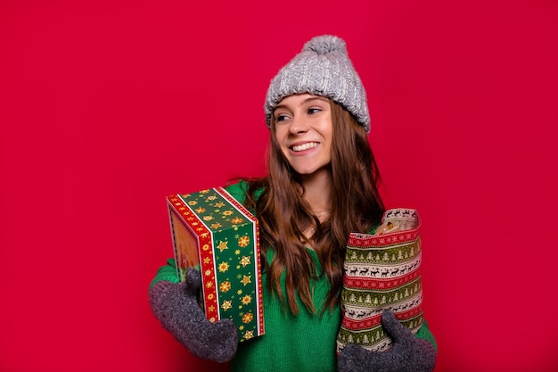 Attractive happy woman with long light-brown hair and wonderful smile dressed winter grey cap, mittens and green sweater holding new year presents and smiling on isolated red background