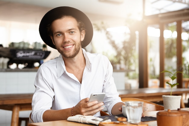 Attractive happy young bearded man in trendy hat texting messages via social networks and browsing internet, using free wifi on his electronic device during coffee break at restaurant