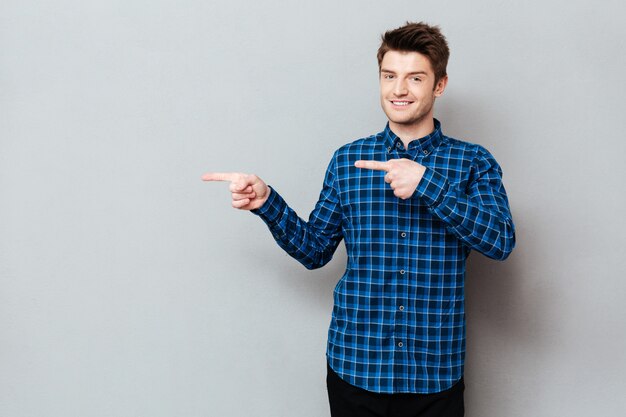 Attractive happy young man standing over grey wall and pointing