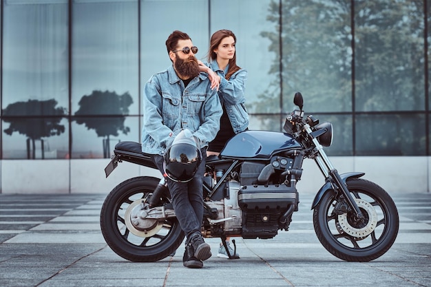 Attractive hipster couple - bearded brutal male in sunglasses and jeans jacket sitting on a retro motorcycle and his young sensual girl standing near, posing against skyscraper.