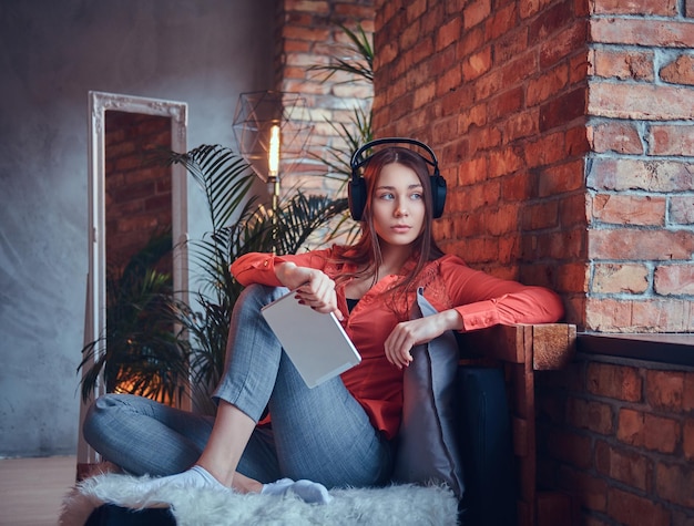 Una ragazza attraente hipster vestita con abiti casual sorride mentre ascolta musica eccitante tramite buone cuffie. rilassarsi in una stanza con interni soppalcati.