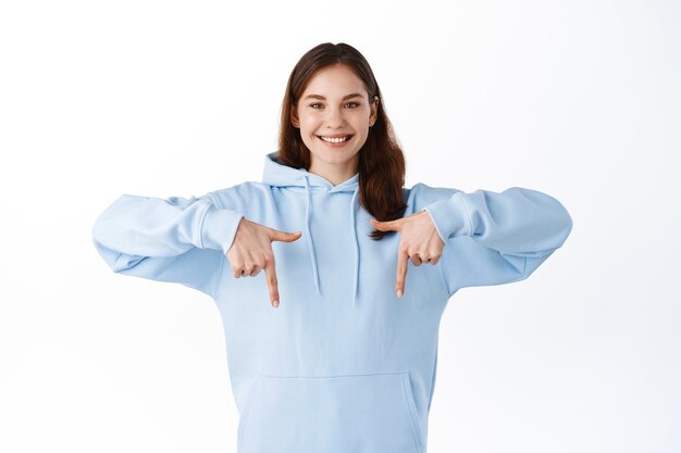 Attractive hipster girl with happy smile showing promotional text advertisement, pointing fingers down at logo, inviting check out promo, standing over white wall