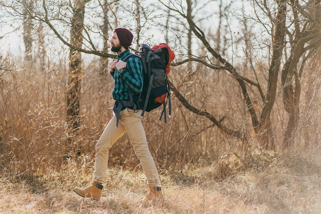 Attractive hipster man traveling with backpack in autumn forest wearing checkered shirt and hat