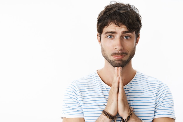 Free photo attractive hot boyfriend making puppy eyes and pouting while holding hands in pray, asking for help, favour or forgiveness making innocent sincere look to persuade womanfriend he sorry over gray wall