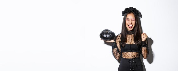 Free photo attractive joyful asian girl enjoying halloween holding black pumpkin and cheering wearing witch cos