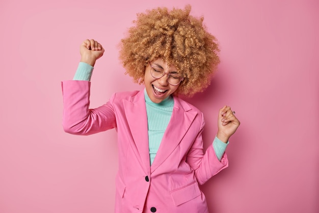 Attraente gioiosa donna dai capelli ricci fa sentire la danza del trionfo come un vincitore vestito con abiti eleganti celebra i risultati personali isolati su sfondo rosa finalmente ho raggiunto il mio obiettivo